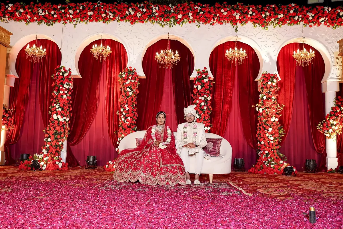 Wedding stage with red velvet arches, red and white flower columns and chandeliers, couple seated on a white sofa, decorated by Mayur Events Bilaspur
